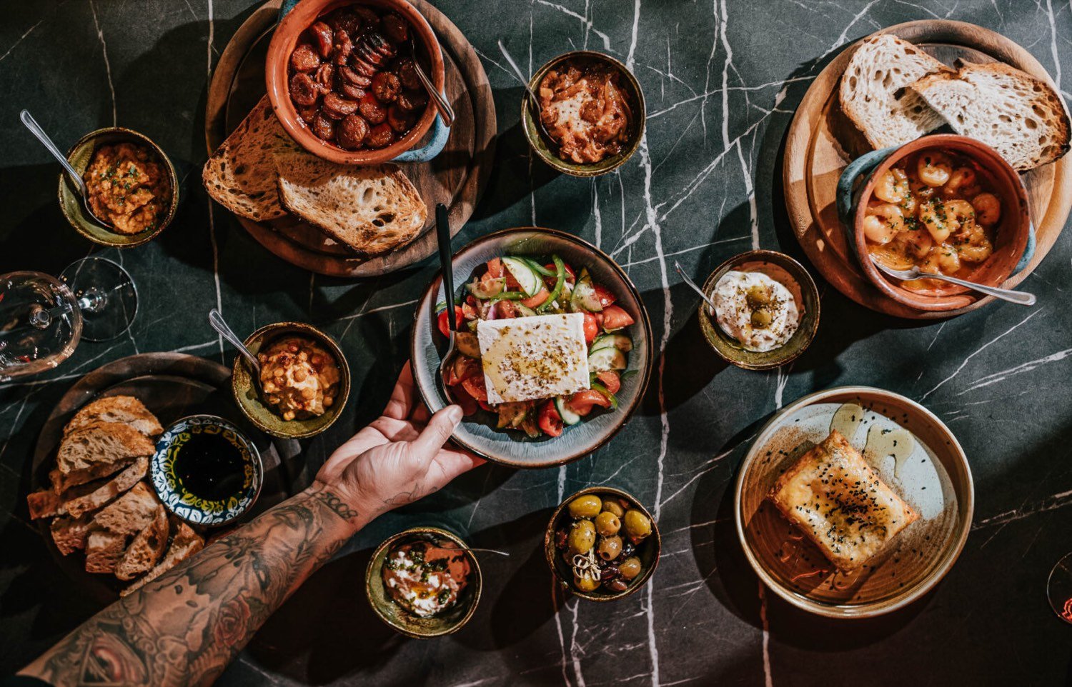 A marble table covered with Mediterranean small plates — chorizo, feta, olives, prawns, hummus and sourdough